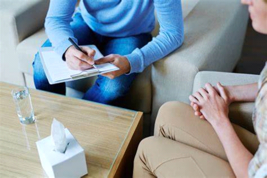 A therapist taking notes on a clipboard while a client sits opposite with hands clasped, in a counselling session