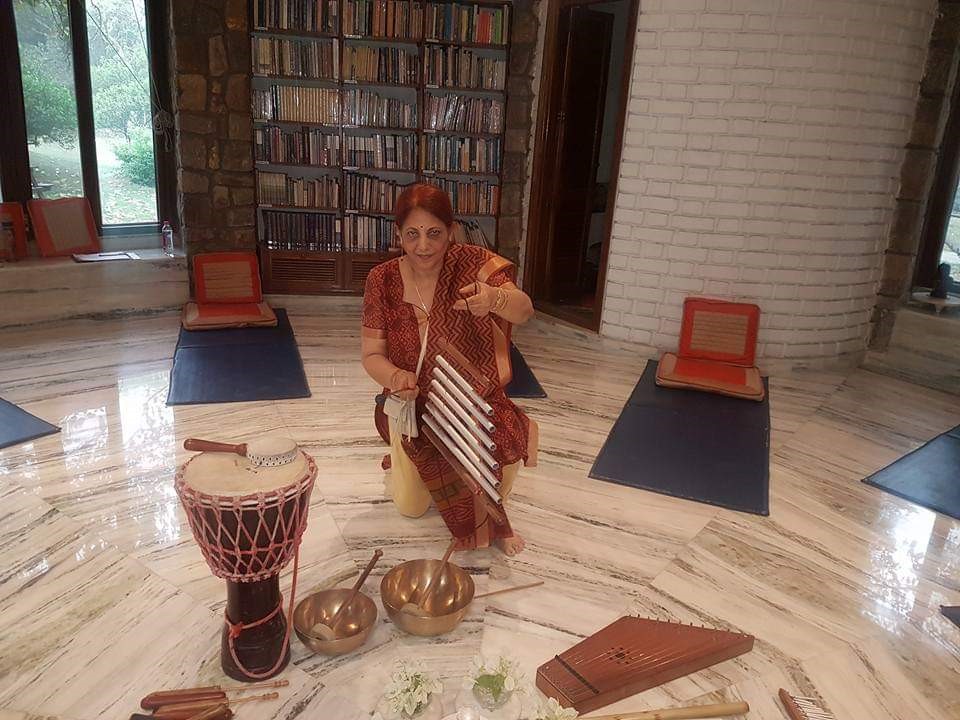 A therapist kneeling beside sound healing instruments, including a djembe drum, singing bowls and chimes, in a session room