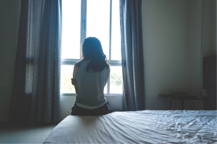 A woman sitting on a bed with her back to the camera, looking out of a window
